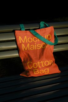 Bright orange tote bag with yellow text, teal handles on metal bench