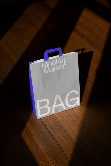 White paper shopping bag mockup with blue handles on a wooden floor with contrasting shadows for product design presentation.