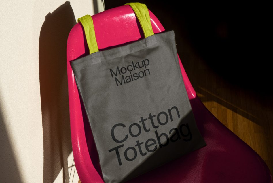 Gray tote bag mockup on a pink chair with natural lighting, showcasing custom text design, perfect for designers to display branding work.