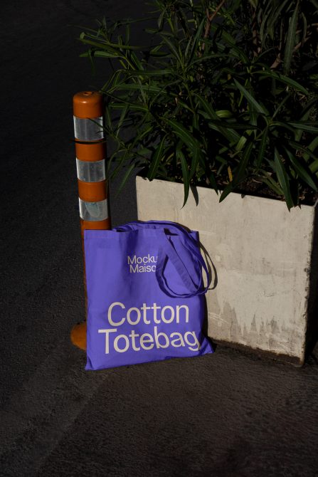 Purple tote bag mockup leaning against an urban planter next to a traffic pole, showcasing custom design space for prints and branding.