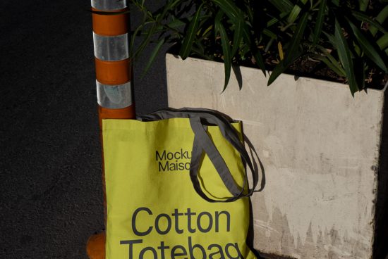 Bright yellow cotton tote bag mockup leaning against a gray planter next to a traffic cone, urban setting for graphic designers.