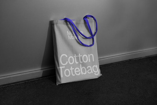 Stylish cotton tote bag mockup with blue handles leaning against a gray wall, ideal for presenting design work to clients.