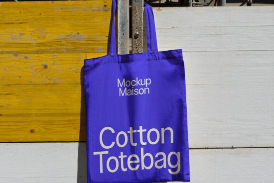 Purple cotton tote bag mockup hanging on a metal hook against a yellow and white wooden background, perfect for showcasing design work.