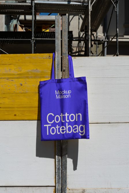 Purple cotton tote bag mockup hanging on metal pole with urban construction background for product design display.