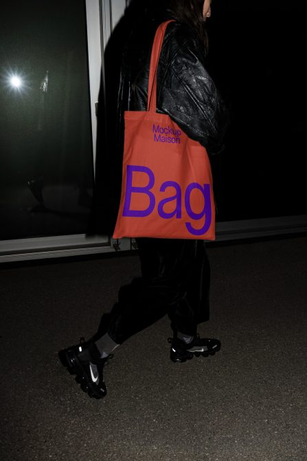 Person side view carrying vibrant orange tote bag mockup with bold typography, leather jacket, casual style, night setting, streetwear fashion.