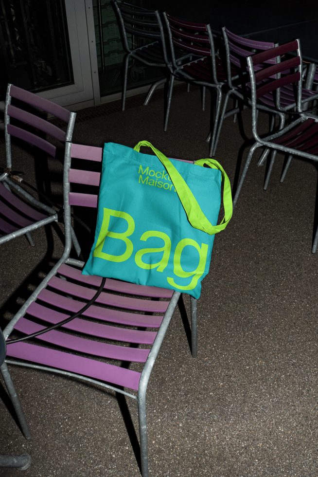 Turquoise tote bag with bold yellow typography on a metal chair, mockup design showcasing bag design against stacked chairs background.