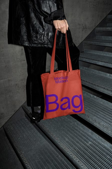 Person holding a red tote bag mockup with blue text, urban setting with concrete stairs, ideal for designers looking for realistic bag mockups.