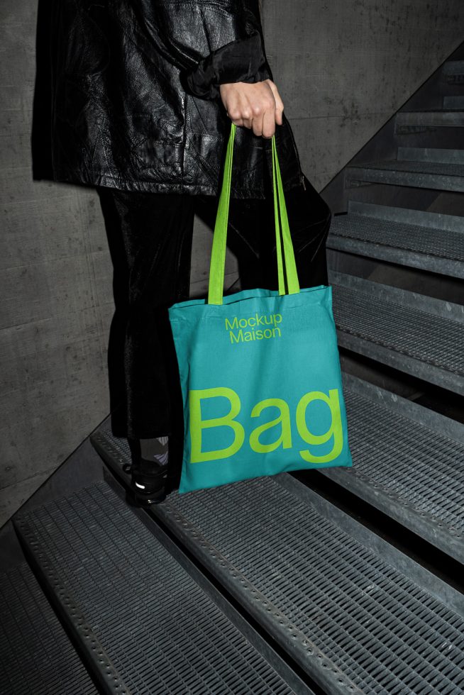 Person holding a teal tote bag mockup with bold yellow text, standing on concrete stairs, ideal for branding presentations in mockups category.