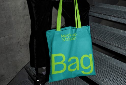 Tote bag mockup held by person on stairs, vibrant turquoise with bold yellow typography, realistic urban setting, perfect for showcasing design work.