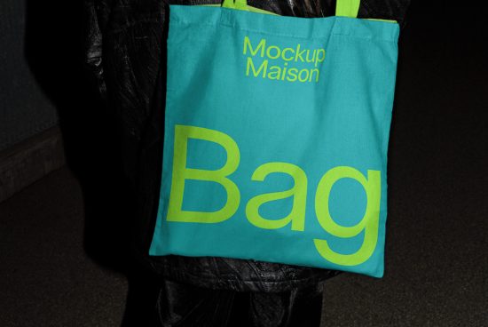 Tote bag mockup with bold typography design on teal fabric held by person in black jacket, ideal for presentations, graphic design assets.