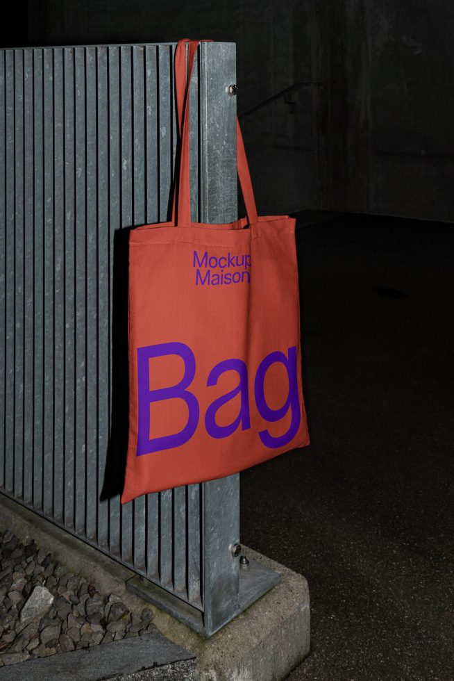 Red tote bag mockup hanging on a metal fence, featuring bold typography, ideal for designers to showcase branding or font designs.