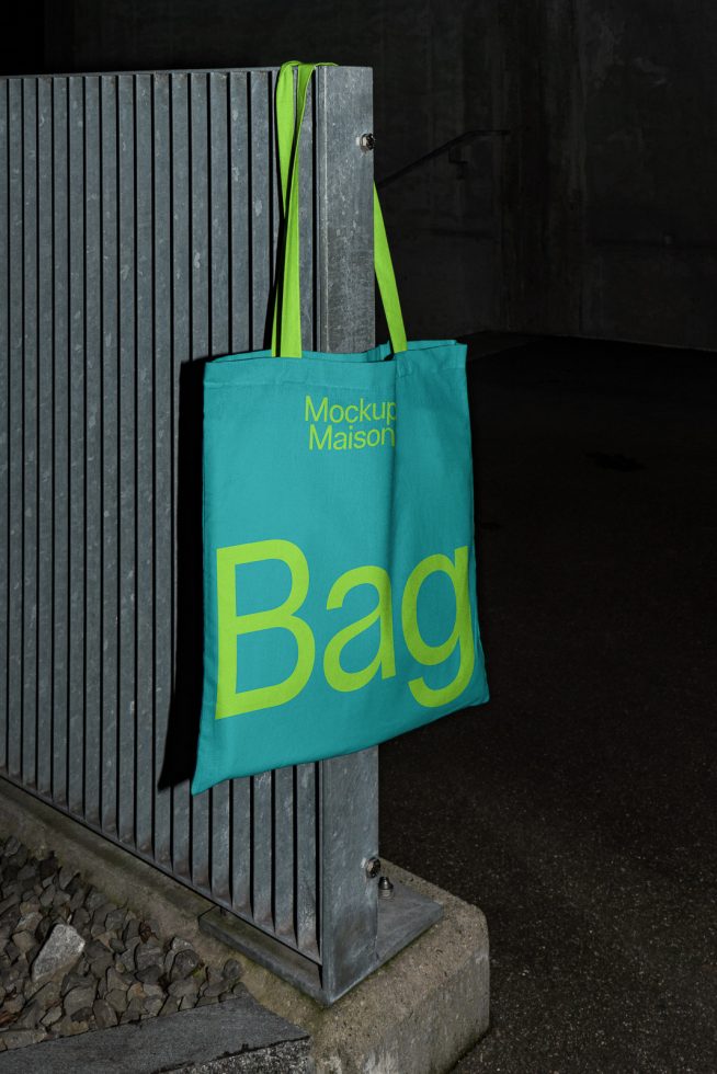 Teal tote bag mockup with bold yellow font hanging on a metal bench in an urban setting, night-time lighting, perfect for branding presentations.