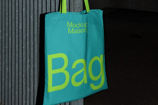 Teal tote bag mockup with yellow bold font, hanging on gray textured background, spotlighting mockup potential for designers.