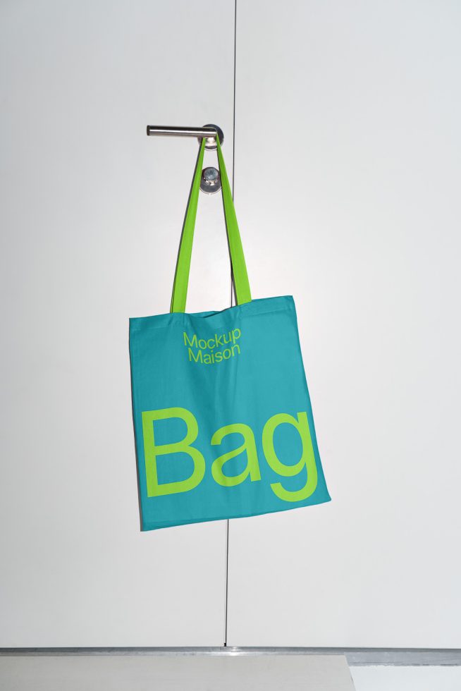 Turquoise tote bag mockup with bold yellow typography hanging on a modern steel hook against a white backdrop suitable for design presentations.