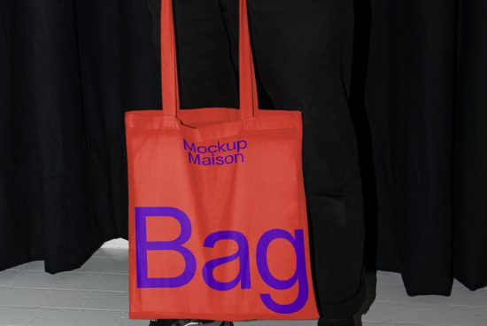 Red tote bag mockup with bold blue text, hanging against a black curtain background, ideal for showcasing design work in a realistic setting.