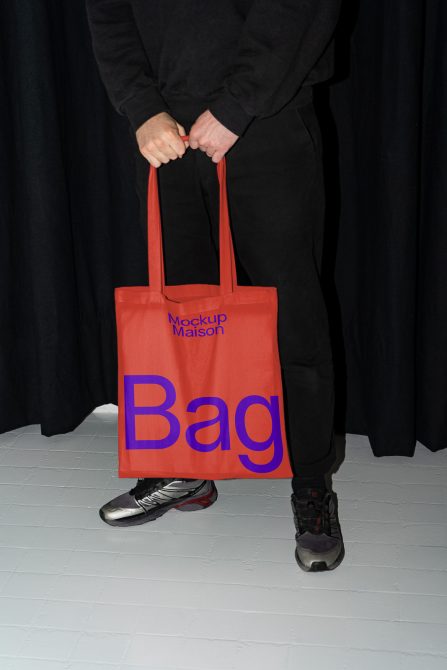 Person holding red tote bag mockup with bold blue text, ideal for design presentations, on black and white background.