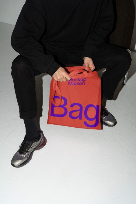 Person holding red tote bag mockup with blue text for design showcase, sitting in dark attire against neutral background for graphic presentation.