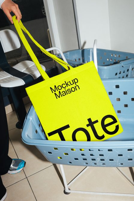 Bright yellow tote bag mockup held by a person, contrasting with a blue shopping basket, ideal for showcasing design work.