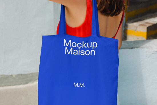 Blue tote bag mockup with white text held by a person in a red top against an urban backdrop, ideal for designers looking for realistic product display.