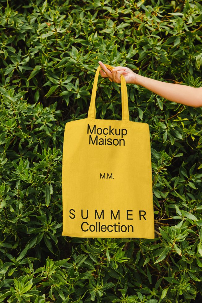 Hand holding yellow tote bag mockup with typography design on green leafy background, ideal for eco-friendly branding presentation.