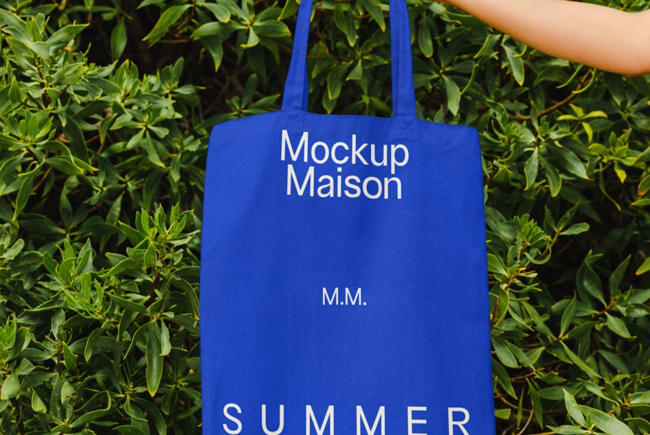 Blue tote bag mockup with white text 'Mockup Maison' held against green foliage background, ideal for eco-friendly design presentations.