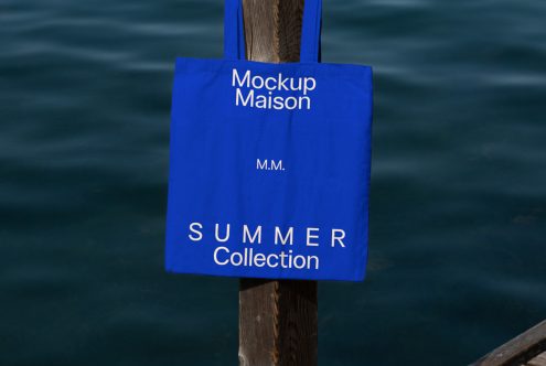 Blue tote bag mockup hanging on wooden post with text Mockup Maison M.M. SUMMER Collection over water backdrop, for graphic presentation.