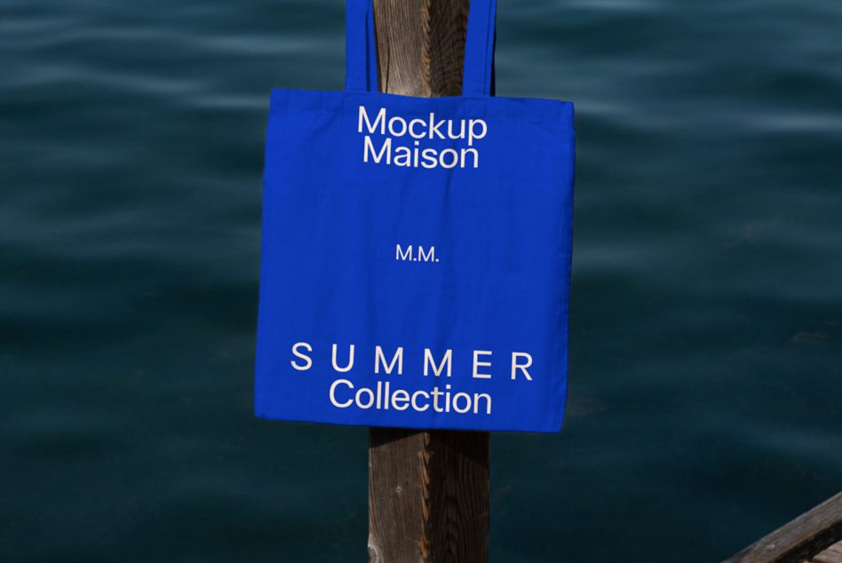 Blue tote bag mockup hanging on wooden post with text Mockup Maison M.M. SUMMER Collection over water backdrop, for graphic presentation.