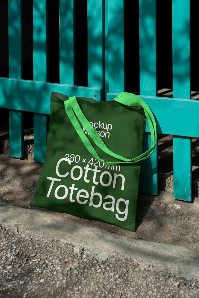 Green cotton tote bag mockup leaning against a blue wooden fence, realistic shadows, design asset for graphic presentations.