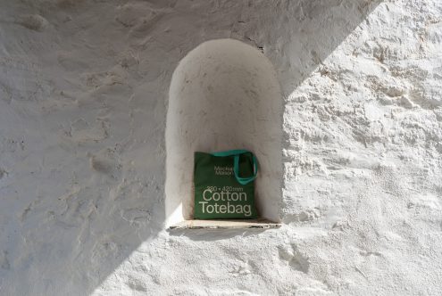 Green cotton tote bag mockup on a rustic white wall in bright sunlight, perfect for showcasing bag designs and logos for designers.