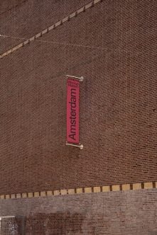 Red vertical banner hanging on brick wall with black Amsterdam text centered