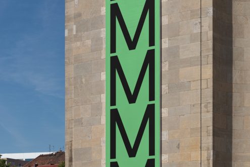 Green banner with bold black 'M' lettering on building facade, clear sky backdrop, modern design graphic poster, urban typography art.