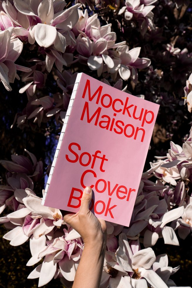 Hand holding a soft cover book mockup against blooming flowers, ideal for book design presentations, realistic digital asset for graphic designers.