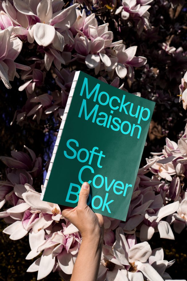 Hand holding a book mockup with a teal cover against a blooming magnolia background, perfect for designers to showcase cover designs.