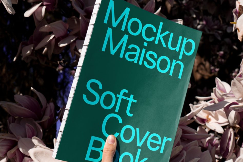 Hand holding teal spiral notebook among flowers showing cyan text Mockup Maison Soft Cover Book