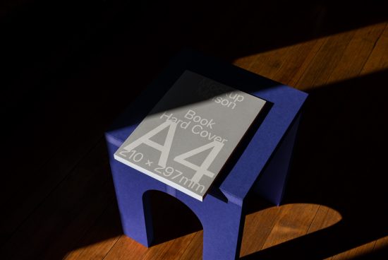 A4 hardcover book mockup on a blue stool with sunlit shadows, elegant presentation for designers, perfect for showcasing cover designs.