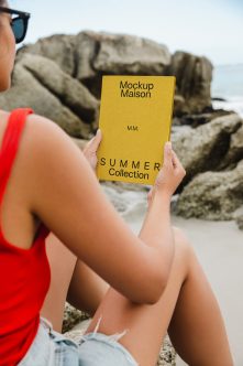 Bright yellow book mockup held at beach with sunglasses, rocks in background