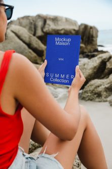 Person in red tank top holding blue hardcover book at beach with boulders