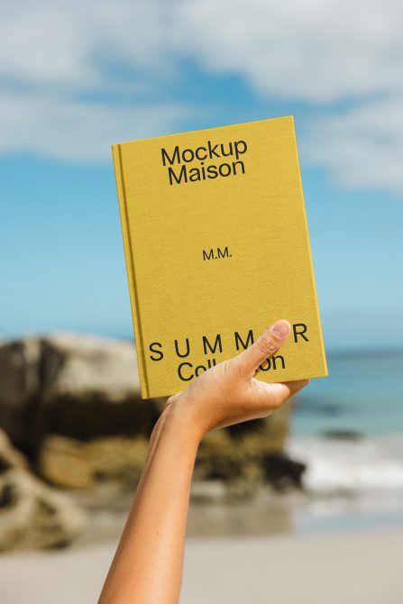 Hand holding yellow book mockup with title "Mockup Maison" on a sunny beach background, perfect for designers presenting summer projects.