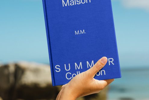 Hand holding a blue book mockup labeled Maison Summer Collection against a blurred beach backdrop, ideal for book cover design presentations.