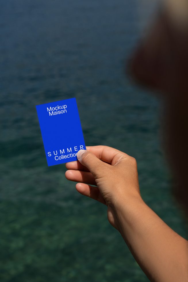 Hand holding blue card with text Mockup Maison Summer Collection against sea background, ideal for designers seeking summer-themed mockup templates.