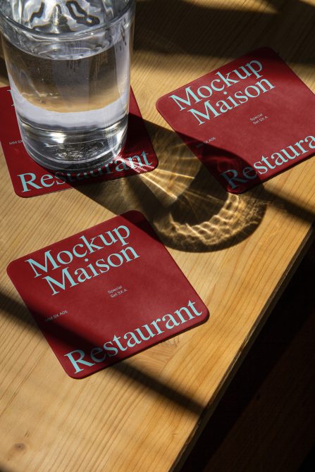 Red coaster mockup with elegant typography lying on a wooden table beside a water glass, showcasing natural light and shadow play ideal for restaurant branding.