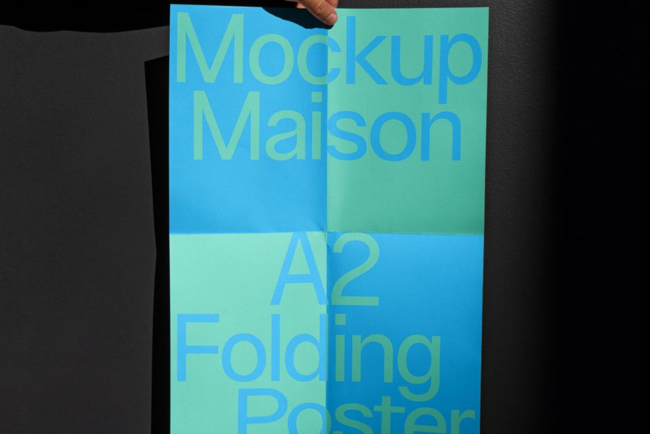 Person holding a blue and green A2 folding poster mockup with typographic design against a black background, graphic design, presentation tool.