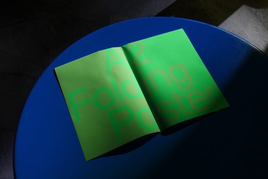A2 folding poster mockup in bright green on blue surface with dramatic lighting and shadow, showcasing print design and typography.