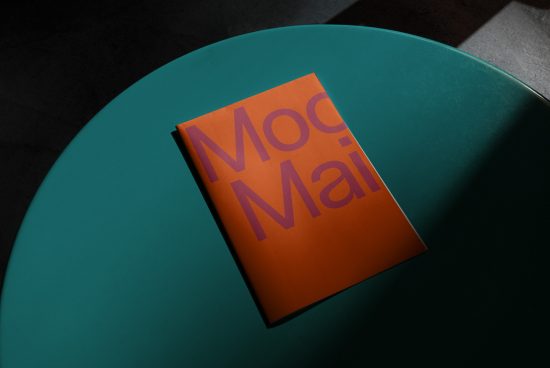 Minimalist magazine mockup with orange cover on teal round table, strong shadow, design presentation.