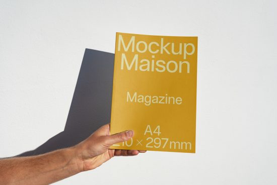 Hand holding a yellow magazine mockup with text Mockup Maison A4 dimensions on a white background with a shadow, clear design mockup presentation.