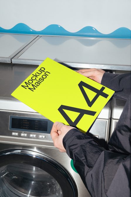 Person holding a bright yellow magazine mockup with bold typography in front of a washing machine, ideal for design presentations.