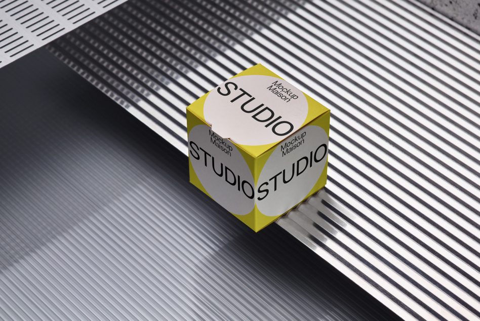 Yellow cube box mockup with rounded white label showing studio branding on striped background