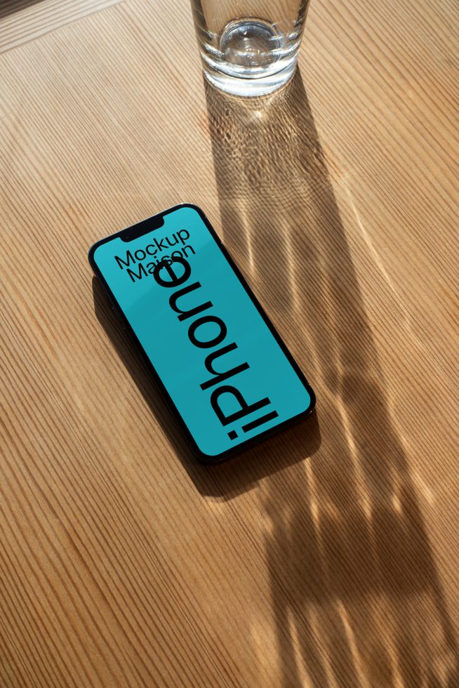 Smartphone mockup on wooden table next to glass of water with light shadows, ideal for presenting app designs and user interfaces in a realistic setting.