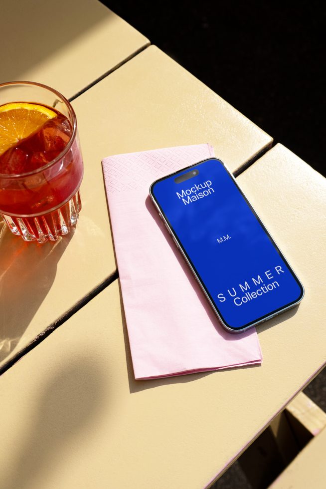 Smartphone mockup on a pink napkin beside a summer cocktail for digital asset designers, perfect for showcasing UI/UX designs.