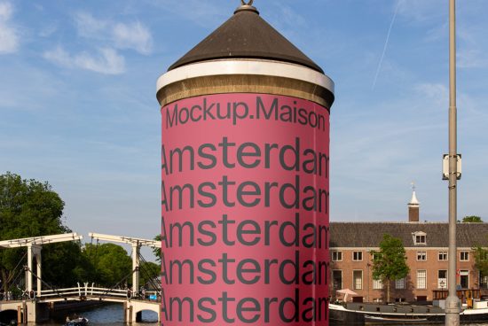Outdoor billboard mockup on cylindrical tower with pink ad space showcasing repetitive 'Amsterdam' text, clear skies and a drawbridge background.